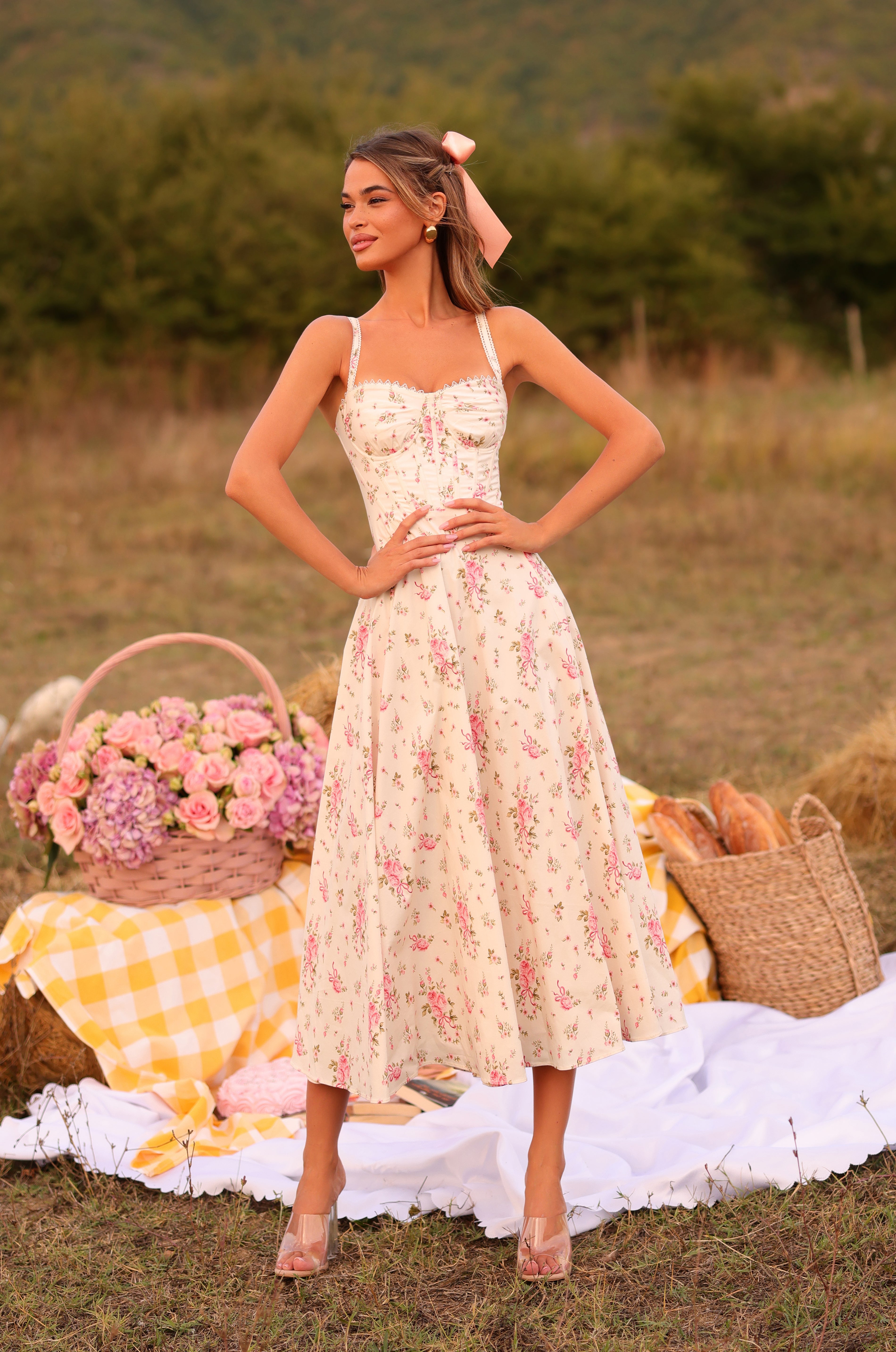 A woman wearing the Cream White Floral Casual Midi Dress: Rosella stands on grass beside a picnic with flowers, bread, and baskets atop a white and yellow blanket, framed by trees and hills in the background—capturing summer style.